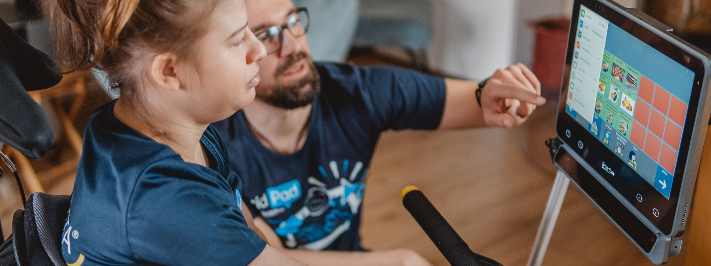 Eine Frau im Rollstuhl sitz vor ihrem Grid Pad. Ein Mann hockt neben ihr und zeigt ihr etwas auf dem Bildschirm