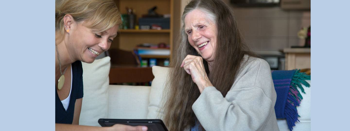 Zwei Frauen sitzen sich gegenüber und lachen. Die jüngere Frau hält ein Kommunikationsgerät in der Hand, das die ältere Frau nutzt, um zu kommunizieren.