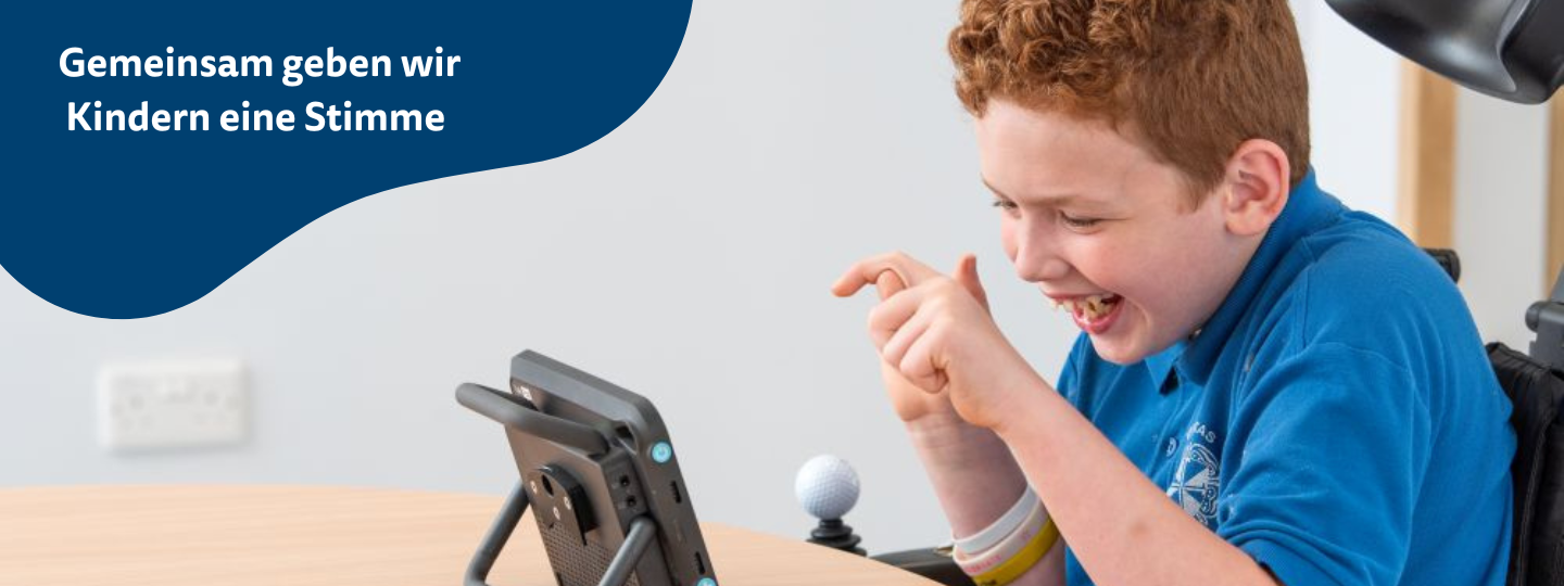 Ein Junge sitzt am Tisch und tippt lachend auf seinen Sprachcomputer.